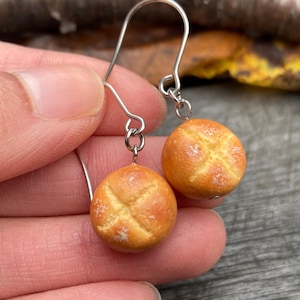 May include: A pair of novelty earrings featuring miniature, round bread rolls with a cross-shaped design on top. The earrings are suspended from silver-colored hooks. The bread rolls are a warm, golden-brown color.