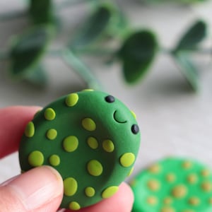 May include: A green, oval polymer clay charm with a smiling face and yellow polka dots. The charm is held in a hand, with a second charm visible in the background. The charm has black eyes and a curved mouth.