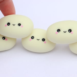 Puede incluir: Colección de cinco mochis ovalados de color amarillo pálido, estilo kawaii. Cada mochi presenta una cara sonriente con ojos negros, boca curvada y mejillas rosadas. Los mochis están dispuestos sobre una superficie blanca.