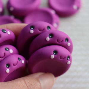 May include: Close-up of several small, magenta clay figures, each with a smiling face, black eyes, and white dots. The figures are clustered together, with some in sharp focus and others blurred. The figures are made of clay.