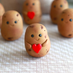 Puede incluir: Un conjunto de figuras caprichosas con forma de patata. Cada patata de color marrón tiene una cara sonriente con ojos azules y un corazón rojo. Las figuras hechas a mano tienen una estética encantadora y juguetona.