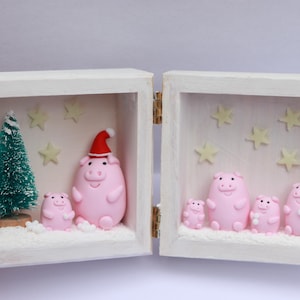 Puede incluir: Una caja de madera blanca con bisagras, con un tema navideño. En el interior, pequeñas figuras de cerdos rosas, un pequeño árbol de Navidad verde y decoraciones en forma de estrella están dispuestas sobre una base nevada. Un cerdo lleva un gorro de Papá Noel.