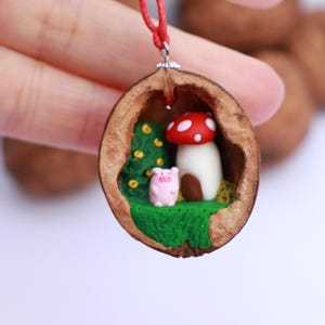 May include: A miniature woodland scene inside a walnut shell, featuring a red and white mushroom house, a pink pig, and green foliage. The walnut shell is attached to a red cord, making it a pendant.