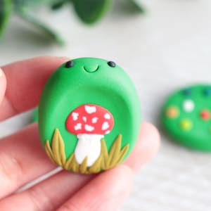 Puede incluir: Un amuleto de arcilla polimérica verde, de forma ovalada, con una cara sonriente, una seta roja y blanca y hierba amarilla. Un segundo amuleto verde con puntos de colores está en el fondo. El amuleto se sostiene en una mano.