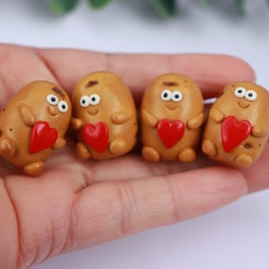 Puede incluir: Cuatro figuras caprichosas con forma de patata, con caras sonrientes y detalles de corazón rojo. Las figuras de color marrón claro tienen ojos blancos y se sostienen en una mano sobre un fondo blanco. Las figuras miden aproximadamente 4 cm de alto.
