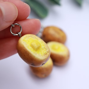 May include: A handmade keychain featuring a miniature, detailed replica of a halved potato with a pat of butter. The potato is brown with a yellow interior, and the butter is a lighter yellow. The keychain is attached to a silver ring.