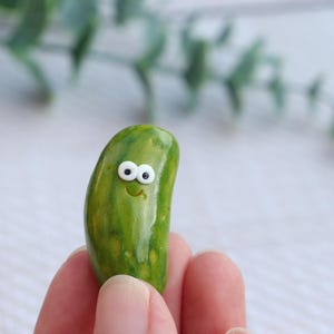 May include: A small, smooth, green pickle figurine with a glossy finish, featuring two white circular eyes with black pupils and a simple etched smile on its front surface, crafted to resemble a miniature vegetable character.