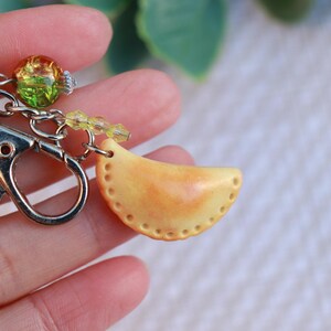 May include: A keychain with a silver clasp and a crescent-shaped charm. The yellow charm has orange shading and small dotted details. It is attached to the clasp with a chain and colourful beads.