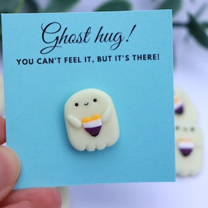 May include: A light blue card with the text "Ghost hug! YOU CAN'T FEEL IT, BUT IT'S THERE!" features a small, off-white ghost pin with a heart-shaped design in yellow, orange, and purple. Additional ghost pins are visible in the background.