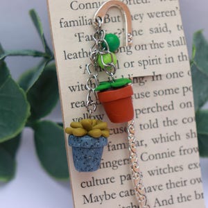 May include: A handmade bookmark with miniature succulent plant charms. The charms include a blue speckled pot with a yellow succulent, and a terracotta pot with a green plant. The bookmark is silver-toned and attached to a book page.