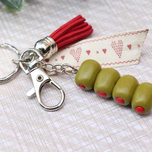 Puede incluir: Un llavero con una argolla y un mosquetón de metal plateado, una borla de ante roja, una cinta de tela color crema con un patrón de corazones rojos bordados y cuatro cuentas verdes en forma de aceituna con centros rojos en fila.