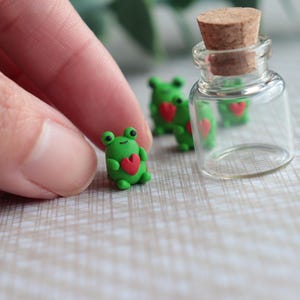 May include: A clear glass bottle with a cork stopper next to several small green frog figurines, each holding a red heart. The frogs are made of clay. The background is a light-coloured surface.