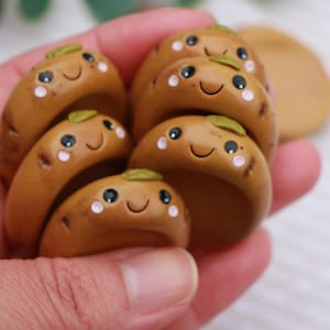 May include: A collection of brown, smiling, potato-shaped clay figurines with green leaf accents. Each figurine has black eyes and white blush spots. The figurines are held in a hand.