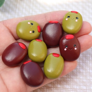 May include: A collection of small, smooth, oval-shaped olive charms in shades of olive green and dark brown. Each piece features a bright red pimento centre and a hand-painted smiling face with black eyes and pink cheeks on the front surface.