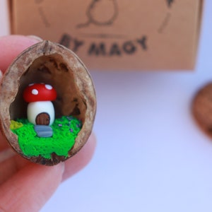 Puede incluir: Una casa de setas en miniatura con un sombrero rojo y blanco se encuentra dentro de una cáscara de nuez partida por la mitad. La cáscara está llena de arcilla verde y marrón, simulando un jardín en miniatura.