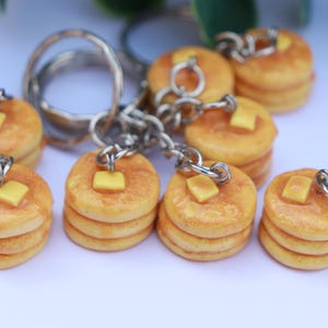 May include: Keyrings with miniature stacks of pancakes, each topped with a pat of butter. The pancakes are golden-brown, and the butter is light yellow. Each keyring has a silver chain and ring.