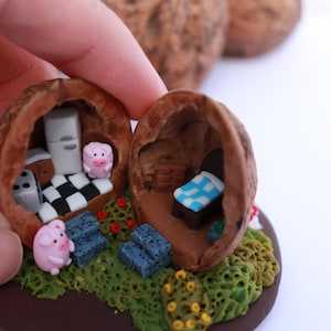 May include: A miniature house crafted from a walnut shell, featuring a pink pig figurine inside. The house is decorated with a black and white chequered floor, a small stove, and a refrigerator. The house is nestled on a bed of green moss.