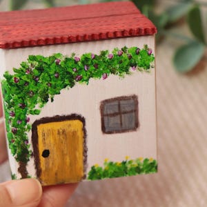 Puede incluir: Una casa en miniatura pintada a mano con tejado rojo, paredes blancas y puerta amarilla. Enredaderas verdes con flores moradas adornan la casa. Una pequeña ventana está pintada en el lateral. Un objeto decorativo encantador.