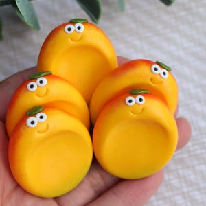 May include: A collection of four small, round, yellow and orange fruit-shaped charms with smooth, concave centres. Each charm features a smiling face with two white eyes and a small green leaf on top, arranged in a cluster to show their design.