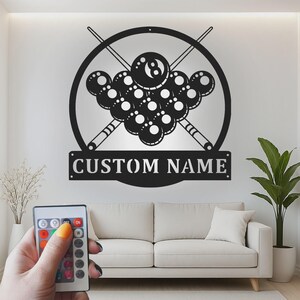 May include: A black metal wall sign featuring a circular frame with two crossed billiard cues and a rack of billiard balls, including an eight ball at the top. Below the circular design is a rectangular banner with the text CUSTOM NAME.