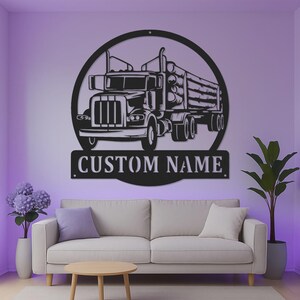 Op de afbeelding: Een zwart metalen wanddecoratie met een cirkelvormig frame dat het gedetailleerde silhouet van een houttruck met boomstammen bevat. Onder de truck bevindt zich een rechthoekige banner met de tekst CUSTOM NAME in hoofdletters in stencilstijl.