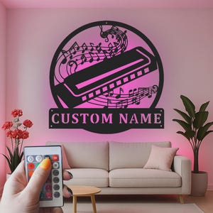May include: A circular black metal wall sign featuring a harmonica silhouette surrounded by musical notes, with a rectangular banner below displaying the text CUSTOM NAME, illuminated by a pink light against a wall above a sofa.