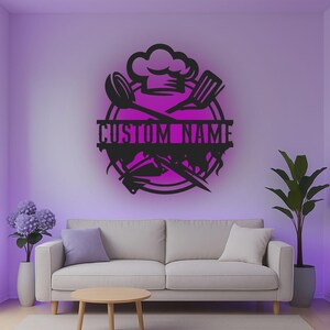 May include: A black circular wall sign featuring a chef hat at the top, crossed kitchen utensils, and a central horizontal bar with the text CUSTOM NAME. The design is mounted on a wall, creating a silhouette effect against a purple background.