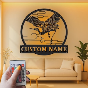 May include: A circular black metal wall art piece featuring a crane in flight against a stylized sun and cloud background. Below the circle is a rectangular plaque with the text CUSTOM NAME in bold, capital letters, illuminated by backlighting.