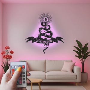 May include: A black wall-mounted sign featuring a coiled snake design with celestial motifs, including stars and moons, topped with a sunburst. A banner across the centre displays the text CUSTOM NAME, with a pink LED glow emanating from behind.