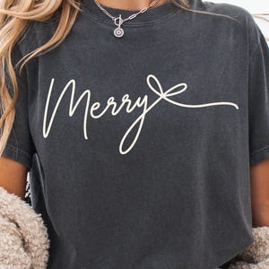 Może przedstawiać: Ciemnoszary t-shirt z kr&oacute;tkim rękawem z napisem Merry wykonanym białą czcionką kursywą. Tekst kończy się stylizowanym rysunkiem liniowym kokardki. Koszulka ma okrągły dekolt i miękki, sprany wygląd materiału.