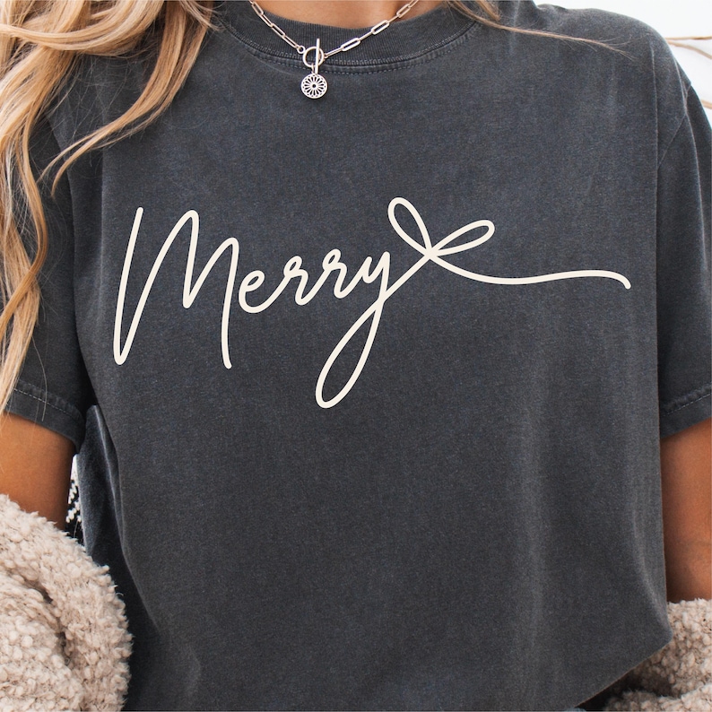 Może przedstawiać: Ciemnoszary t-shirt z kr&oacute;tkim rękawem z napisem Merry wykonanym białą czcionką kursywą. Tekst kończy się stylizowanym rysunkiem liniowym kokardki. Koszulka ma okrągły dekolt i miękki, sprany wygląd materiału.