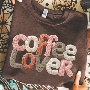 Może przedstawiać: Brązowa bluza z napisem "Coffee Lover" w dzianinowym, trójwymiarowym stylu. Litery są w odcieniach różu, beżu i brązu. Bluza jest złożona i trzymana przez dłonie.
