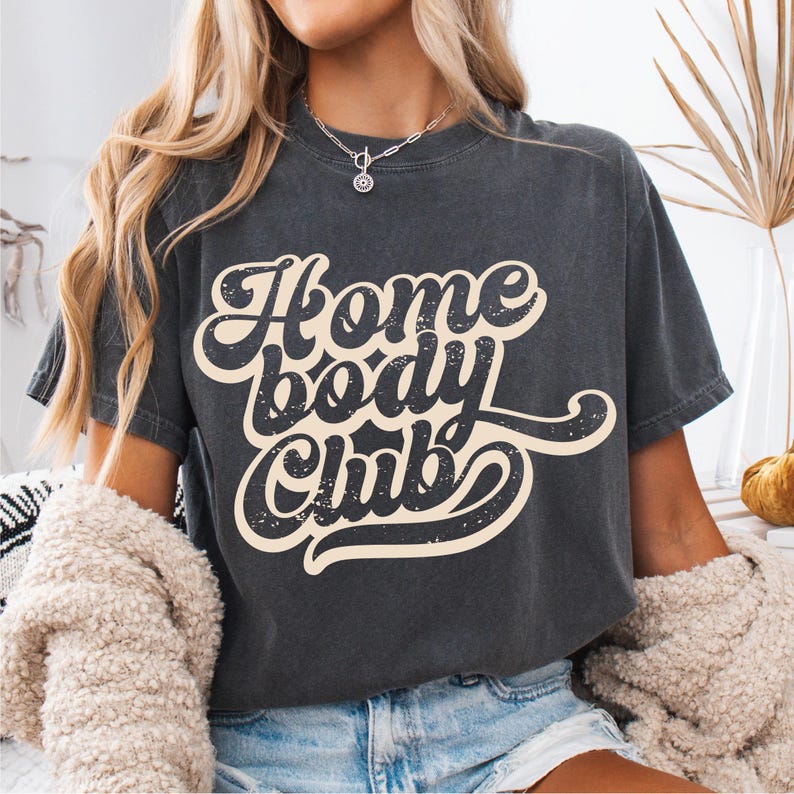 Peut inclure: T-shirt gris fonc&eacute; avec l'inscription "Home Body Club" dans une police r&eacute;tro. Le texte est de couleur cr&egrave;me avec un effet vieilli. Le t-shirt a des manches courtes et un col rond.