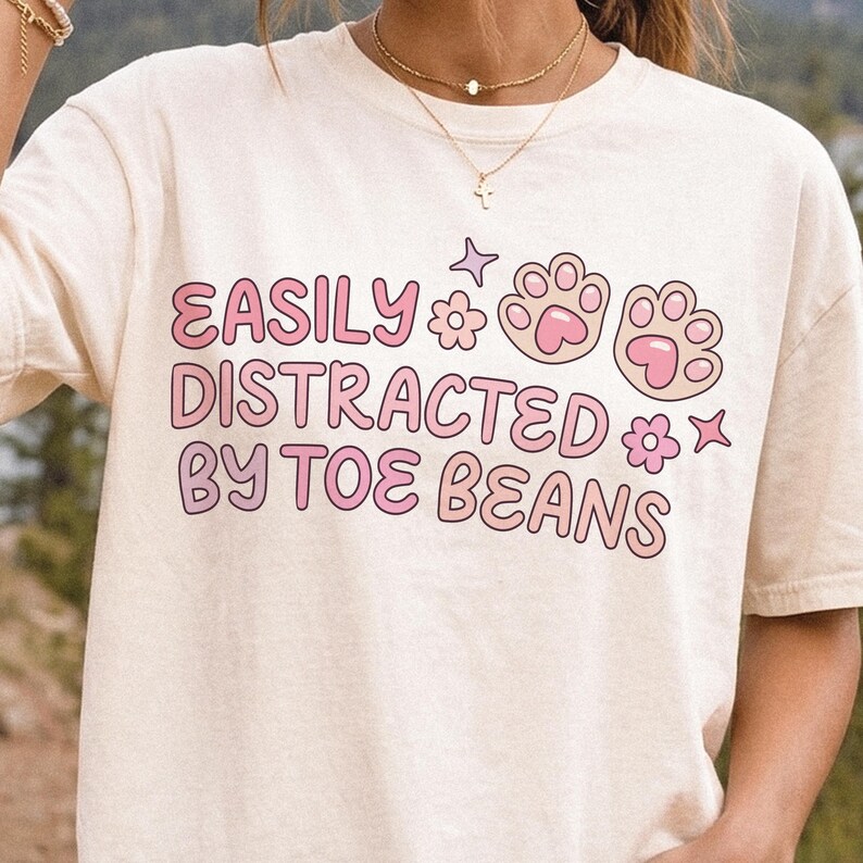 May include: A cream-colored short-sleeve t-shirt featuring the pink text "EASILY DISTRACTED BY TOE BEANS" alongside graphic illustrations of two cat paw prints, two small flowers, and two four-pointed stars printed on the front chest area.