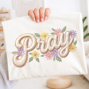 May include: Cream-colored fabric with the word "Pray" embroidered in a textured, off-white font, outlined with a rope-like design. The word is surrounded by colorful embroidered flowers in shades of yellow, pink, and purple, with green leaves.