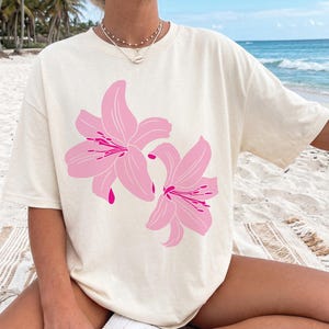 May include: A cream-coloured t-shirt with a large pink floral design. The graphic shows two stylised lilies with detailed petals and vibrant pink accents. The shirt has a relaxed fit, suitable for casual wear.