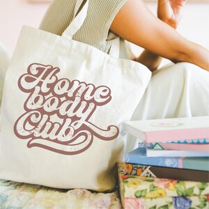 Peut inclure: Sac fourre-tout cr&egrave;me avec l'inscription "Home Body Club" dans une police r&eacute;tro. &Agrave; c&ocirc;t&eacute;, des livres aux couvertures florales et pastel. Le sac a de longues anses et un design simple.