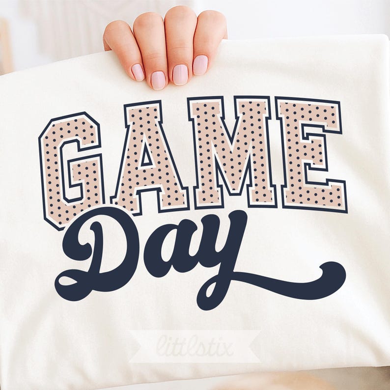 Op de afbeelding: Een wit t-shirt met de tekst GAME DAY. Het woord GAME is geschreven in een groot, blokkerig varsity-lettertype met een beige achtergrond en zwarte stippen. Het woord DAY staat eronder in een donker marineblauw cursief lettertype.