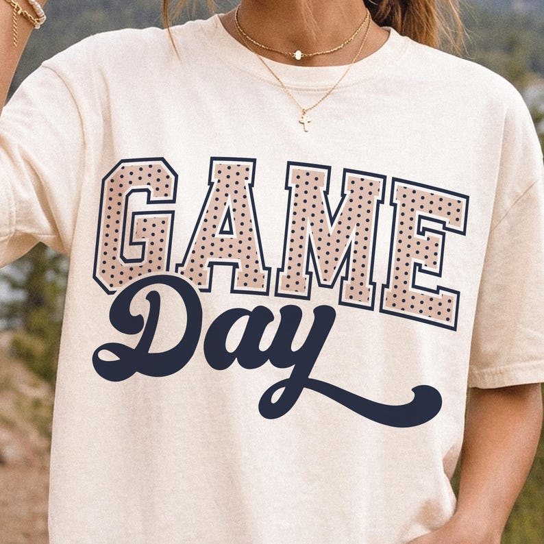 Op de afbeelding: Een cr&egrave;mekleurig T-shirt met korte mouwen en de tekst GAME DAY op de voorkant. Het woord GAME staat in een groot, blokkerig, beige lettertype met zwarte stippen en een donkerblauwe omlijning. Het woord Day is in een donkerblauw cursief lettertype.