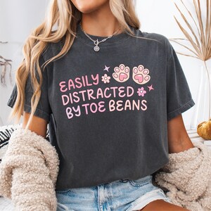 May include: A charcoal gray short-sleeve t-shirt featuring the pink text EASILY DISTRACTED BY TOE BEANS, accompanied by two pink and light pink paw prints and two small flower icons, with a crew neckline and a relaxed, vintage-washed fabric finish.