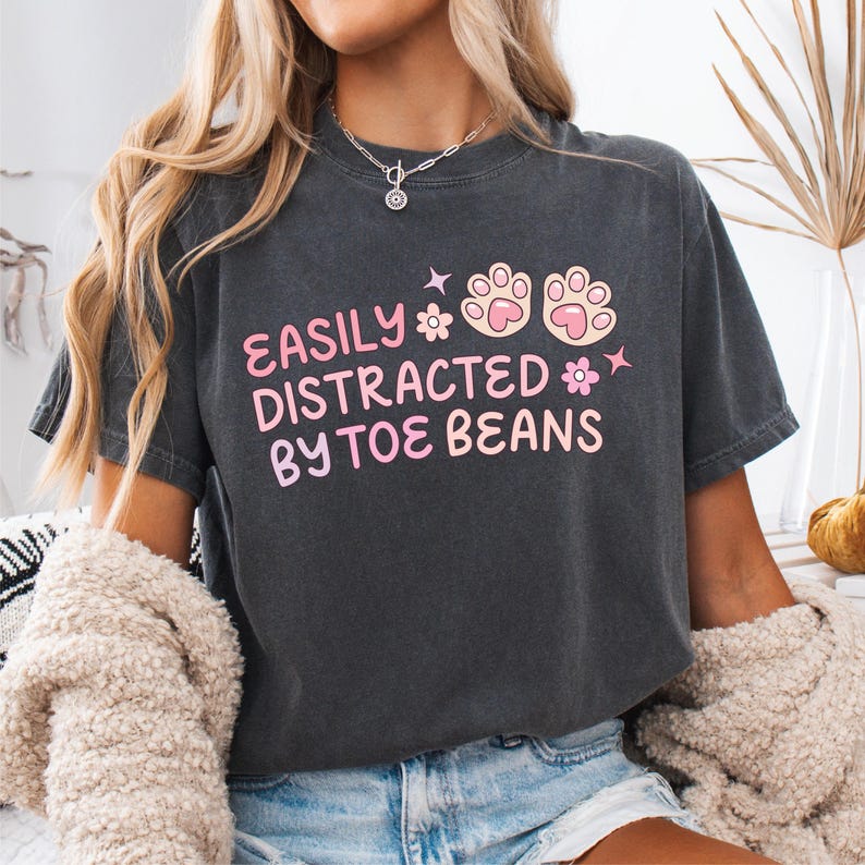 May include: A charcoal gray short-sleeve t-shirt featuring the pink text EASILY DISTRACTED BY TOE BEANS, accompanied by two pink and light pink paw prints and two small flower icons, with a crew neckline and a relaxed, vintage-washed fabric finish.
