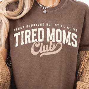 Puede incluir: Una camiseta marr&oacute;n con el texto "SLEEP DEPRIVED BUT STILL ALIVE TIRED MOMS Club" en letras blancas y rosas. La camiseta tiene cuello redondo y mangas cortas. Una mujer lleva la camiseta.