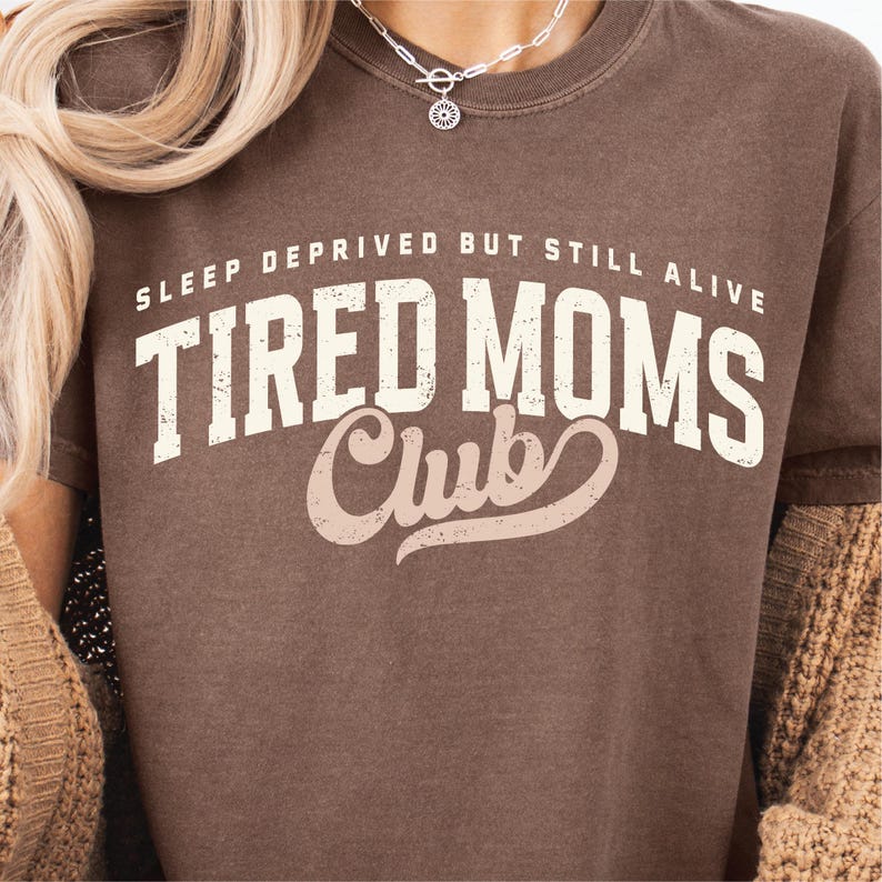Puede incluir: Una camiseta marr&oacute;n con el texto "SLEEP DEPRIVED BUT STILL ALIVE TIRED MOMS Club" en letras blancas y rosas. La camiseta tiene cuello redondo y mangas cortas. Una mujer lleva la camiseta.
