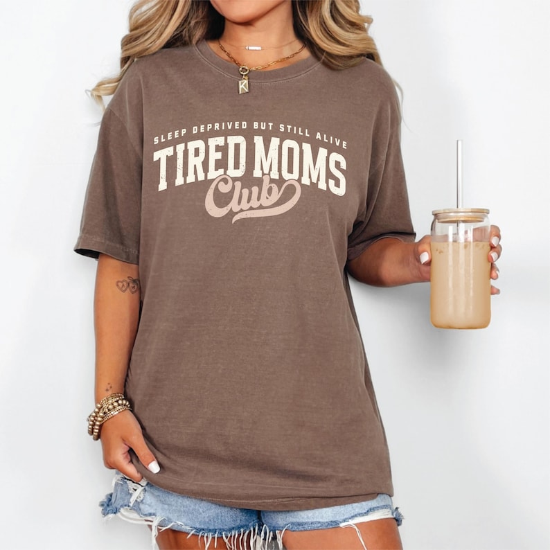 Puede incluir: Una camiseta marr&oacute;n con el texto "SLEEP DEPRIVED BUT STILL ALIVE TIRED MOMS Club" en letras blancas y rosas. La camiseta se usa con pantalones cortos de mezclilla. Se sostiene en la mano un vaso transparente con una pajita y una bebida beige.