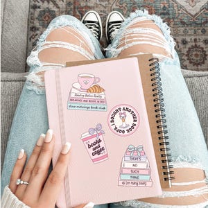 May include: A pink journal with stickers that say "Reading Before Reality", "Breakfast and Books in Bed", "Slow Mornings Book Club", "Books and Coffee", and "I Bought Another Book". A brown spiral notebook is next to it.