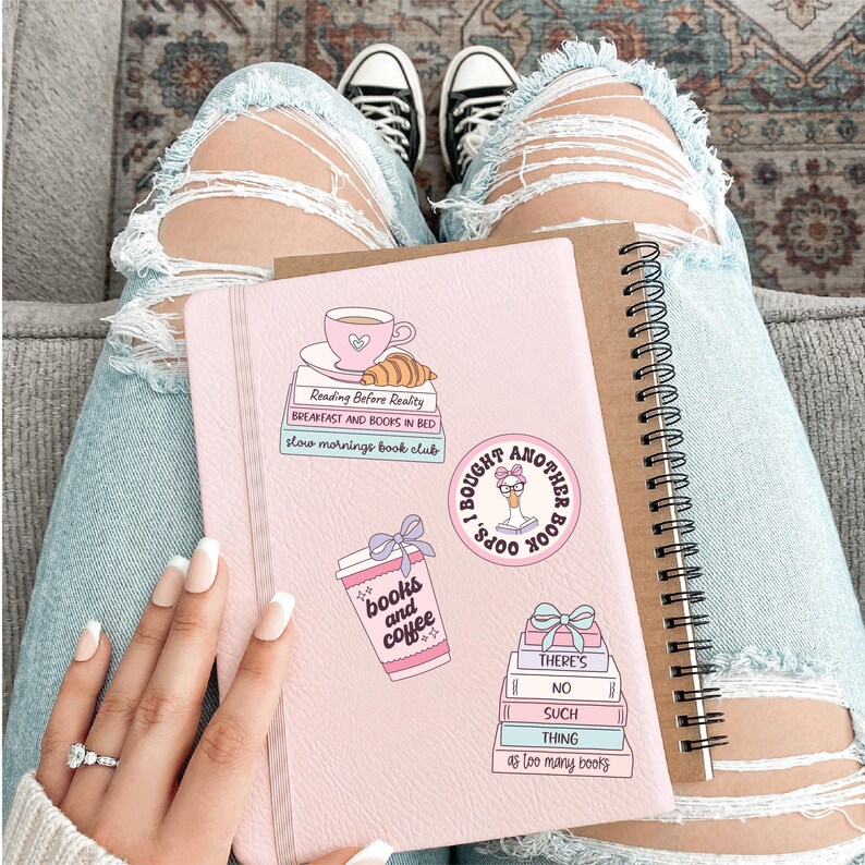 May include: A pink journal with stickers that say "Reading Before Reality", "Breakfast and Books in Bed", "Slow Mornings Book Club", "Books and Coffee", and "I Bought Another Book". A brown spiral notebook is next to it.