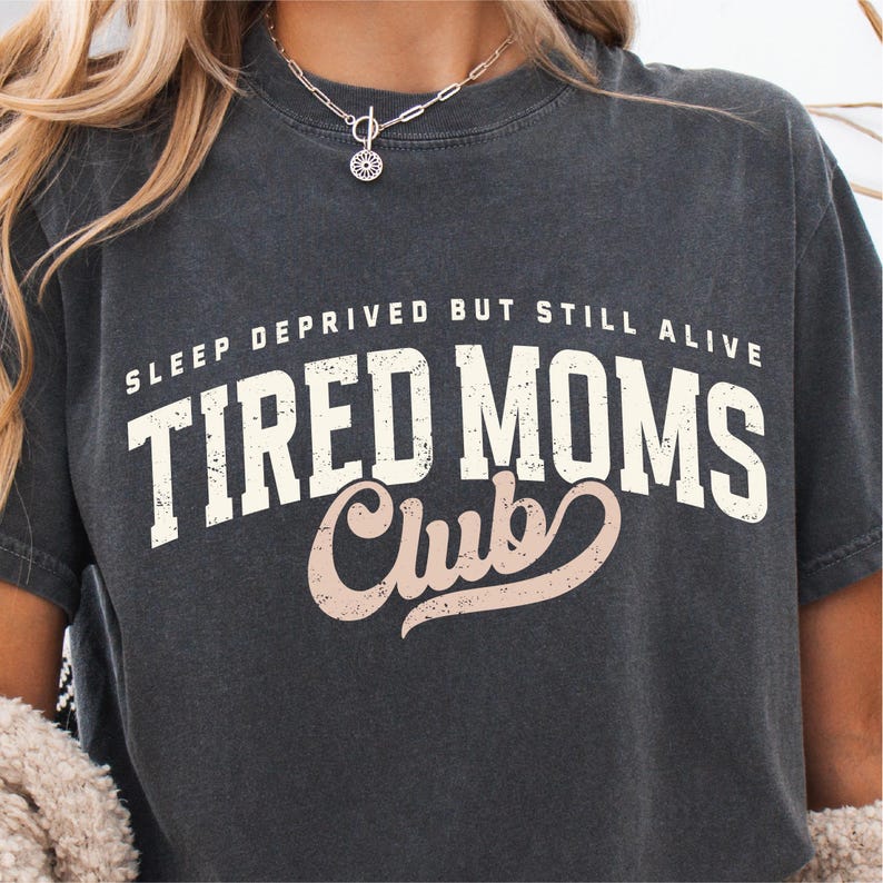 Puede incluir: Camiseta gris oscuro con el texto "SLEEP DEPRIVED BUT STILL ALIVE TIRED MOMS Club" en letras blancas y marr&oacute;n claro. La camiseta tiene un aspecto vintage y desgastado.