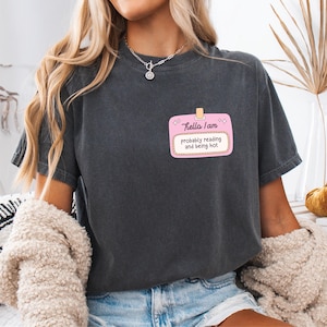 May include: Dark gray t-shirt with a pink name tag graphic that reads "Hello I am probably reading and being hot." The shirt is a casual style, suitable for everyday wear.