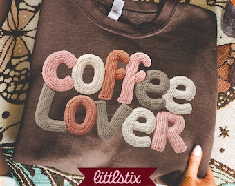May include: A dark brown crewneck sweatshirt featuring the text coffee lover in a textured, knit-style graphic. The letters are rendered in various shades of pink, cream, and tan, mimicking the appearance of yarn. The brand name littlestix is printed in white on a dark red banner at the bottom.
