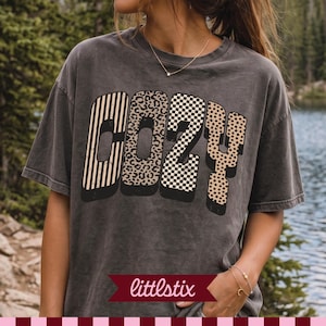 Puede incluir: Una camiseta de manga corta gris carbón con la palabra COZY impresa en letras grandes. Cada letra tiene un patrón distinto: rayas verticales, estampado de leopardo, un tablero de ajedrez en blanco y negro y lunares beige sobre fondo oscuro.